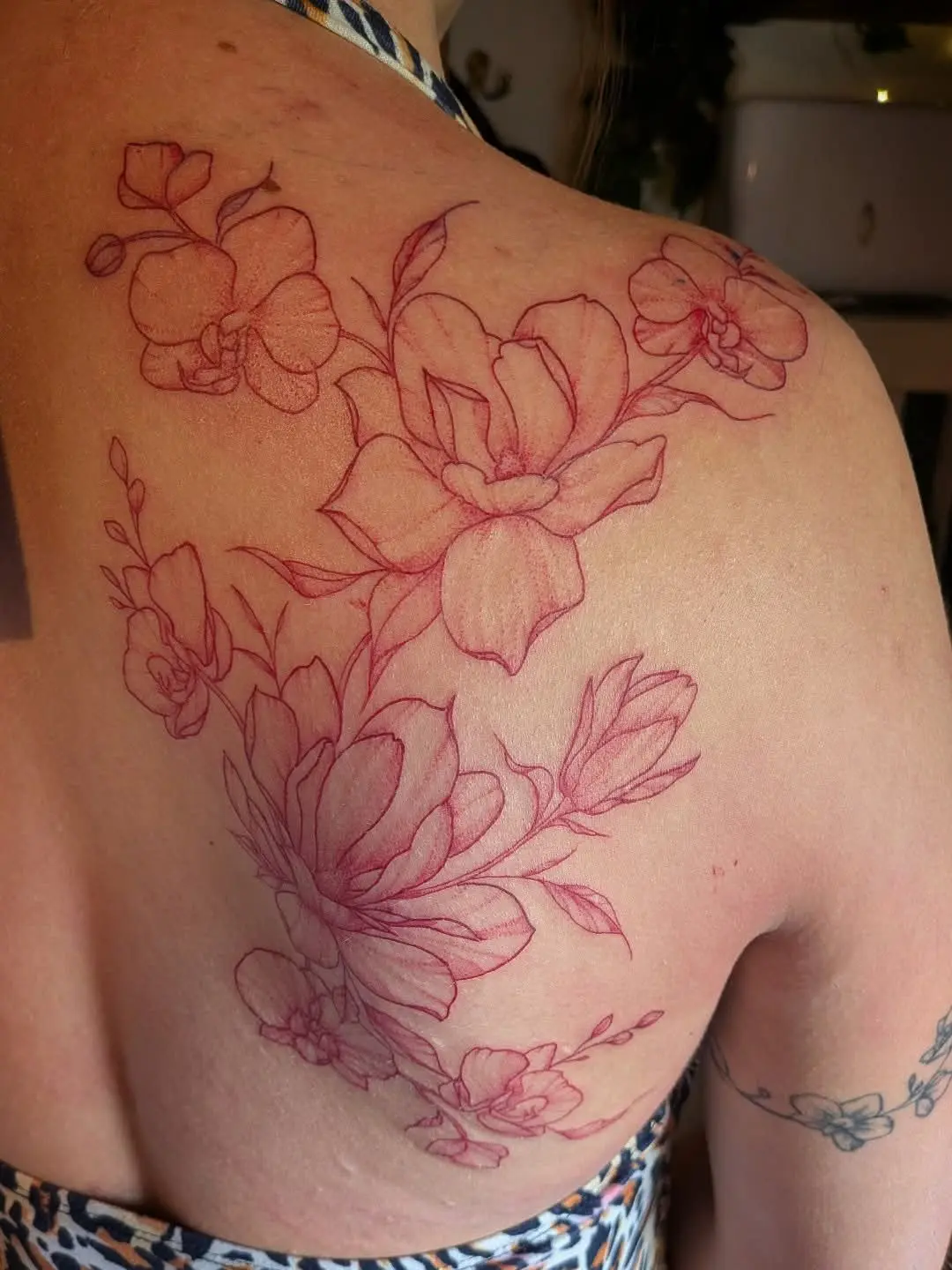 Fine-line red ink tattoo of magnolias and orchids across a shoulder