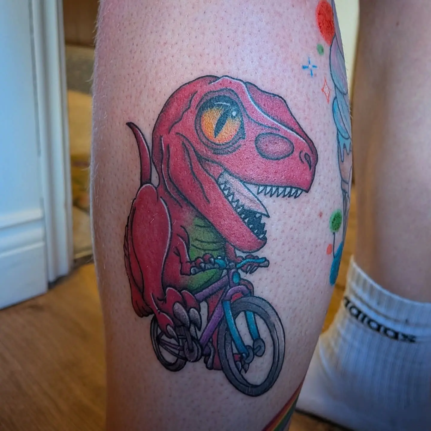Playful colour tattoo of a red dinosaur riding a bicycle
