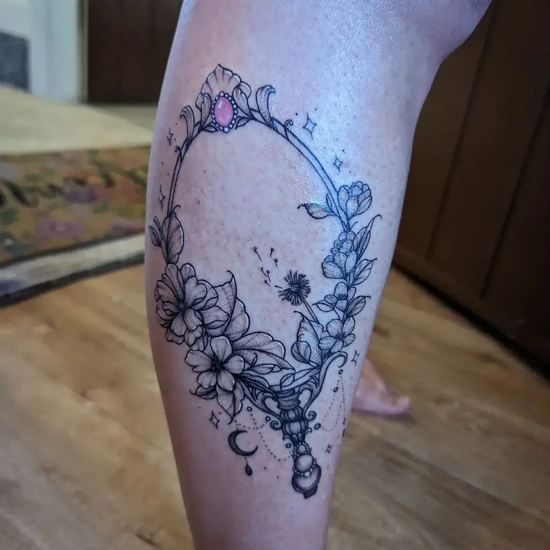 Fine-line tattoo of an ornate mirror wreathed in flowers, with a dandelion and moon