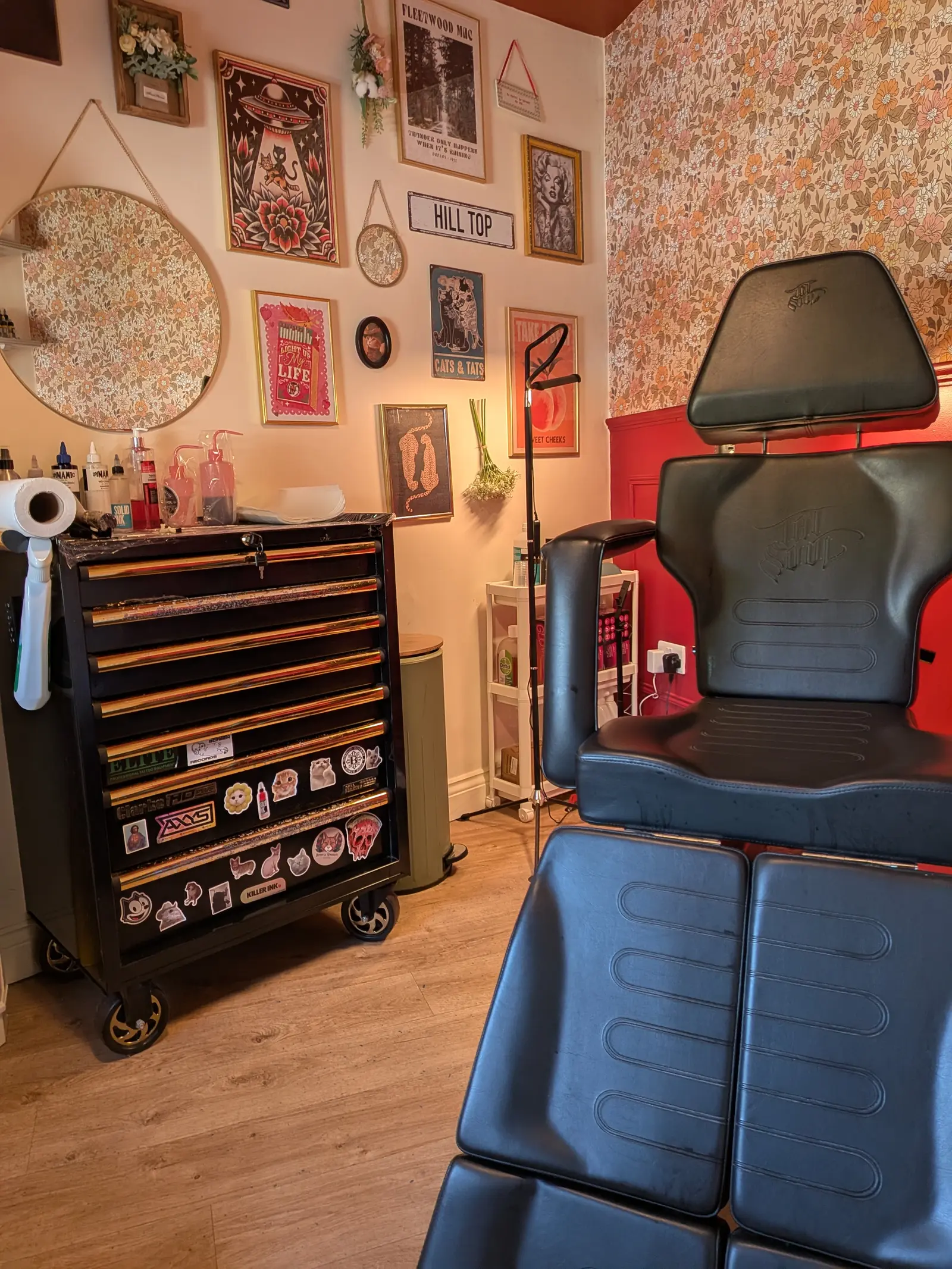 The tattoo chair beside the workstation drawers, under the studio’s gallery wall and floral wallpaper